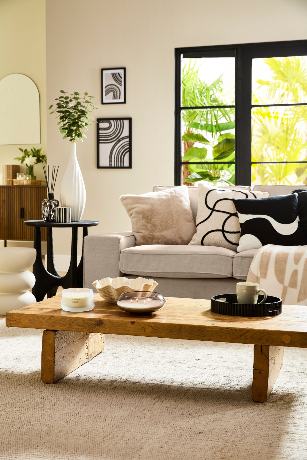 Neutral living room with sofa, patterned cushions, wooden table holding vases and candles, jute rug and window with garden view 