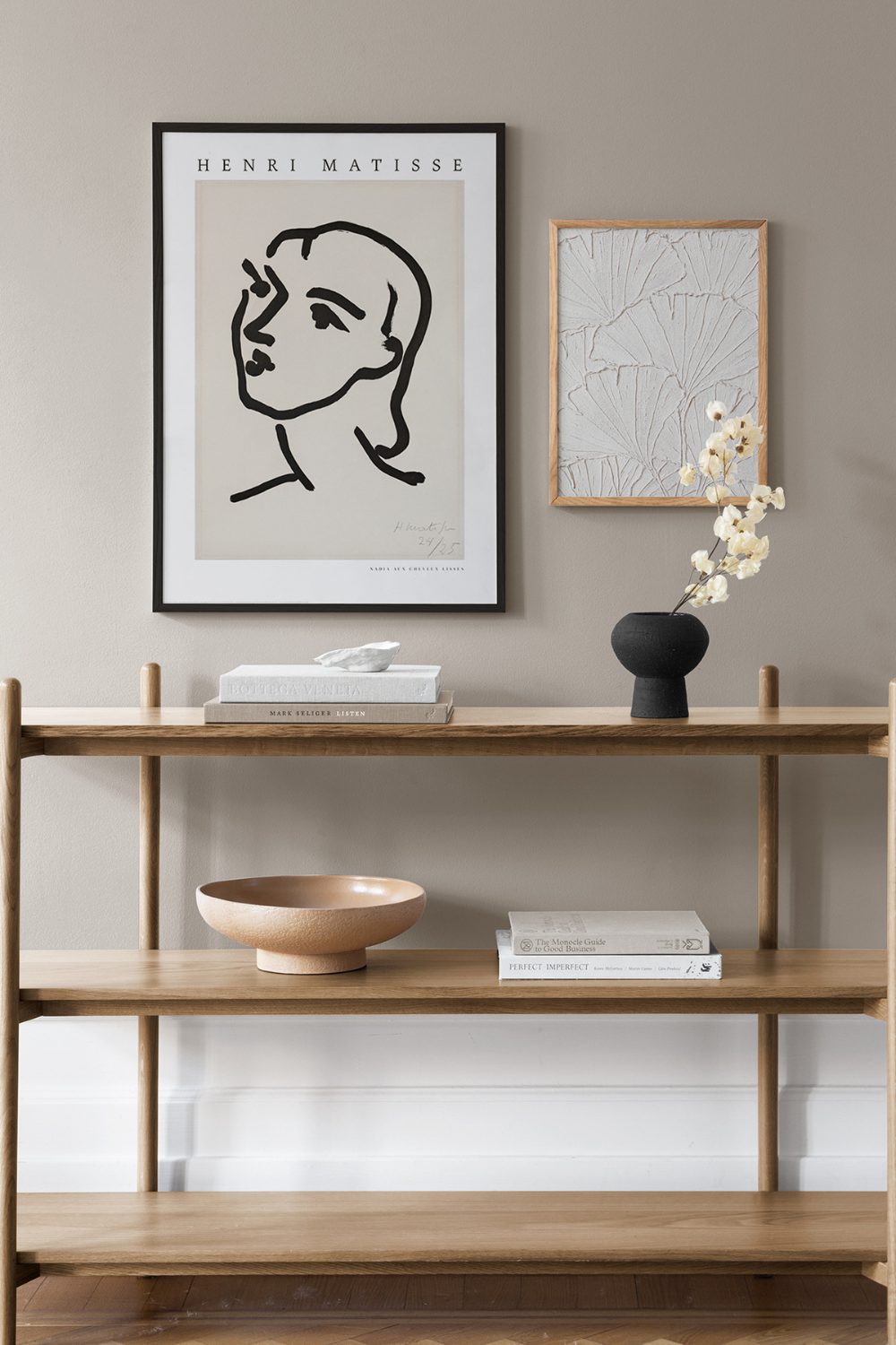 Wooden shelving unit holding books and vases against white wall with framed black and white prints