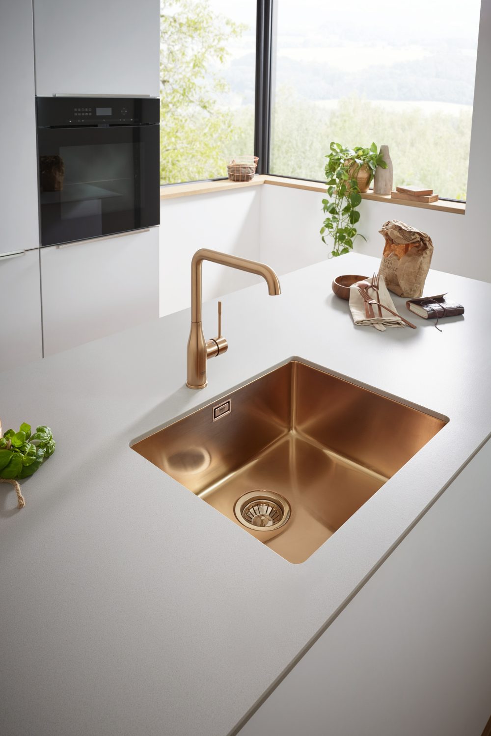 Copper sink in white kitchen counter by corner window with green landscape view