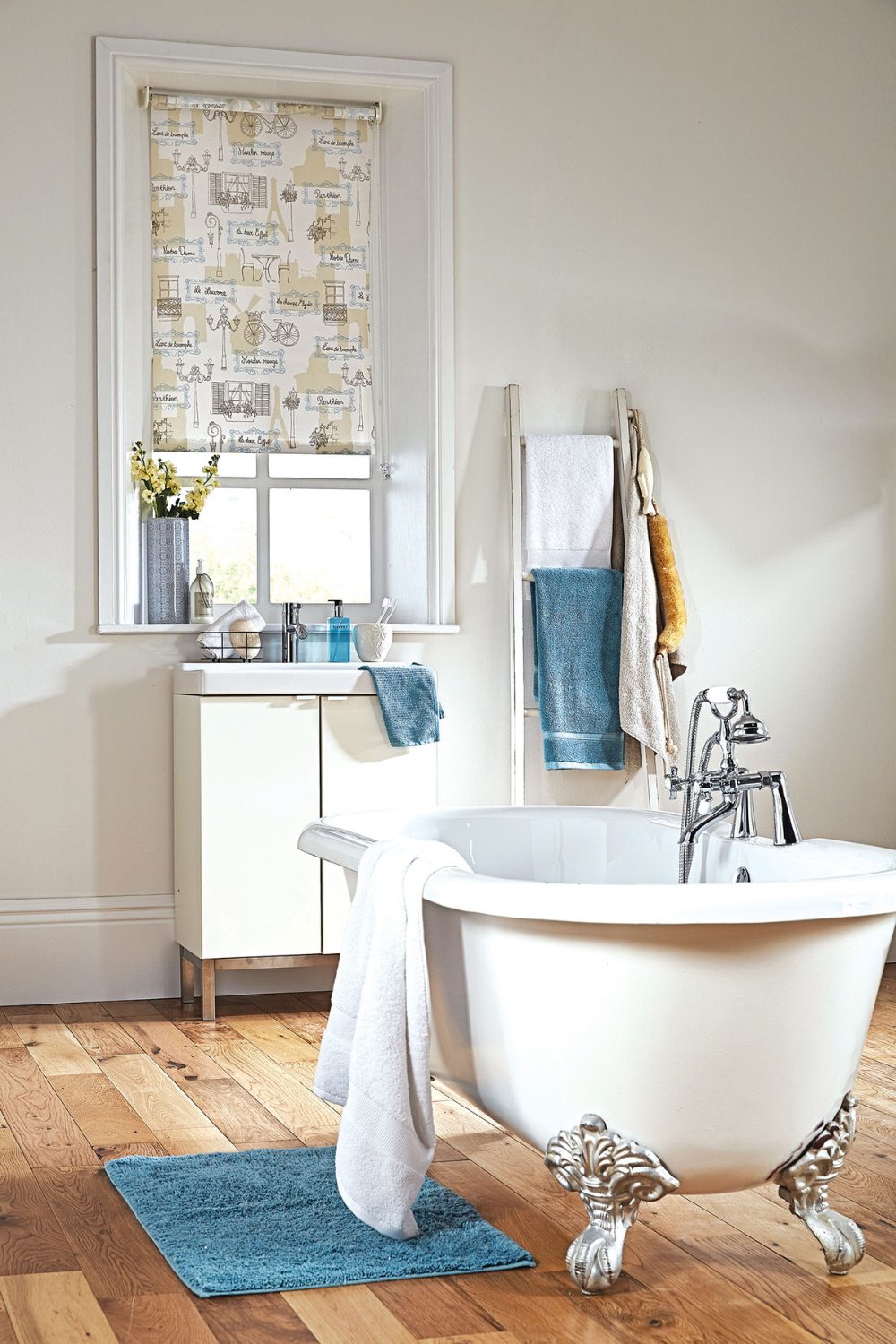 White free-standing bath with silver claw feet on wooden floor with white cabinet, blue bathmat and window with printed blind