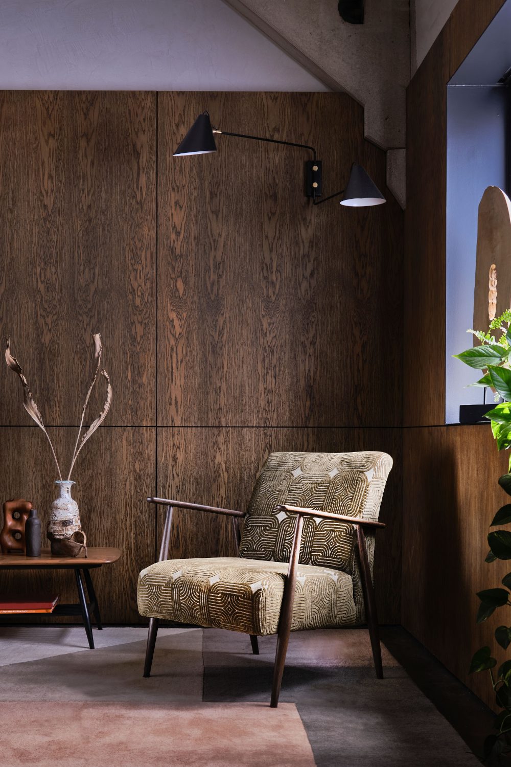 Patterned armchair with wooden arms next to wooden table holding vases against a wooden panelled wall