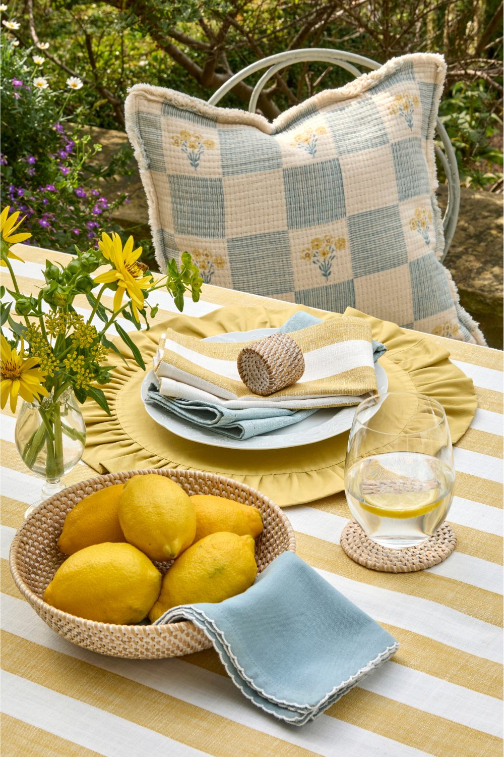 Table covered with yellow & white striped tablecloth holding wicker bowl of lemons, vase of yellow flowers, and blue napkins