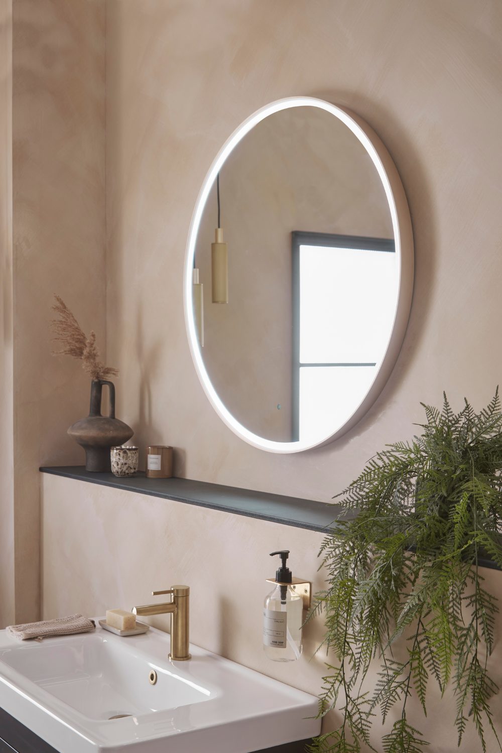 Round mirror on neutral bathroom wall above shelf with candle and pot plant, and white sink