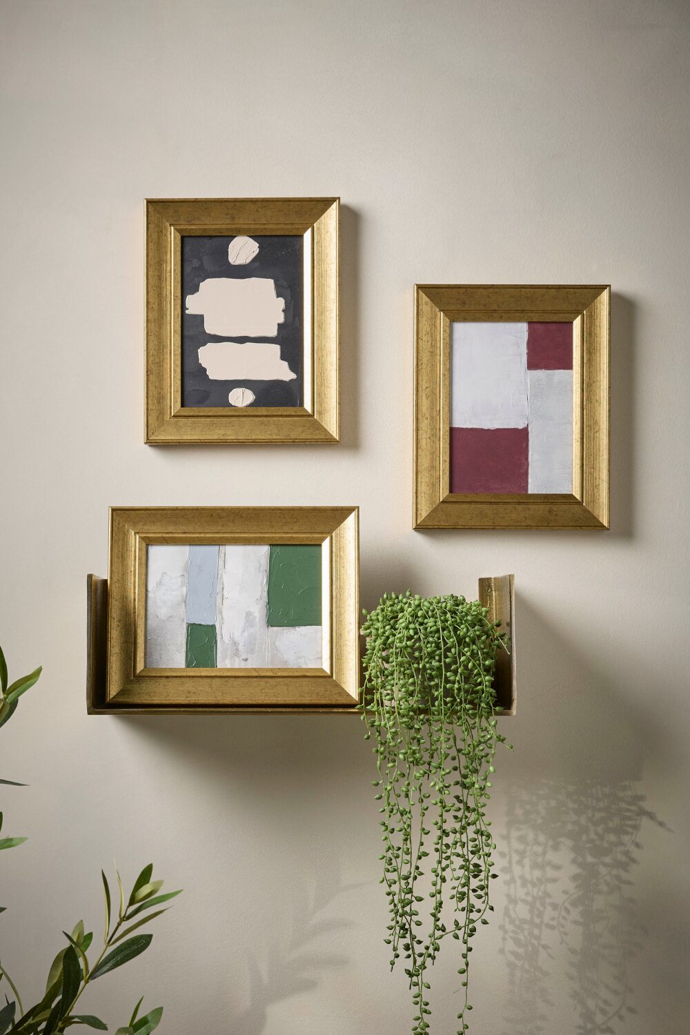 White wall with three gold-framed abstract canvases and shelf with plant pot