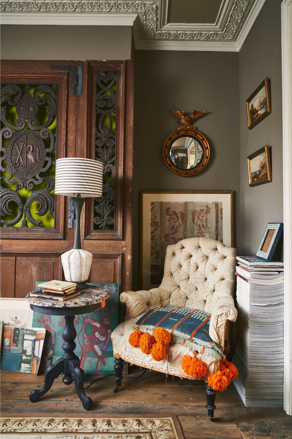 Grey living room with wooden and metal door, round mirror, white armchair, stack of magazines and wooden floor.