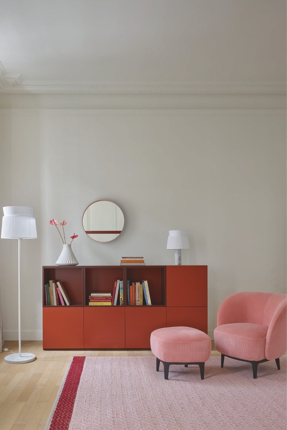 White living room with red sideboard, white lamp, circular mirror, pink armchair and pouffe, and pink rug