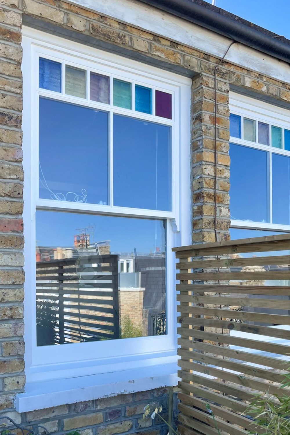 Exterior of a brick house with wooden fence by white sash windows with coloured glass section