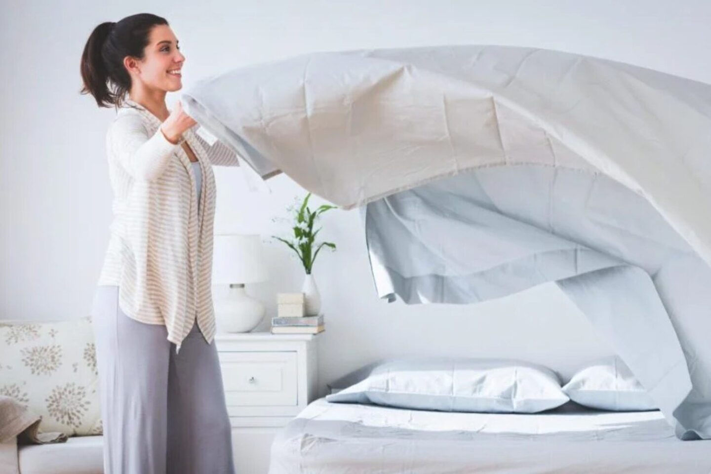 Women lifting a white duvet over a made bed with white bedside table in background