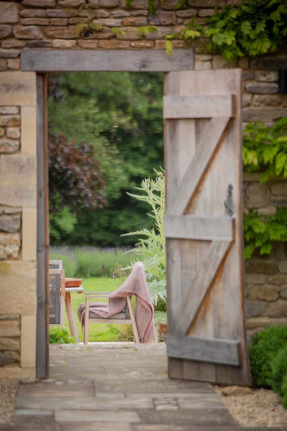 View through a wooden door in stone wall of a wooden table and chairs on green lawn