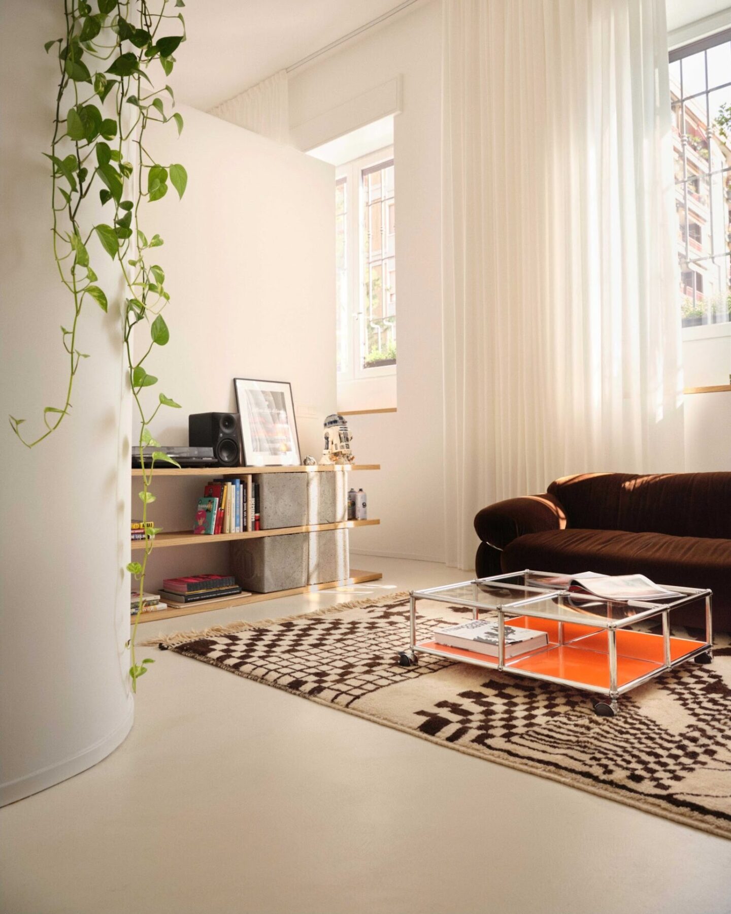 Light bright living room with white walls and curtains, brown sofa, low wooden bookcase and neutral coloured rug with coffee table.