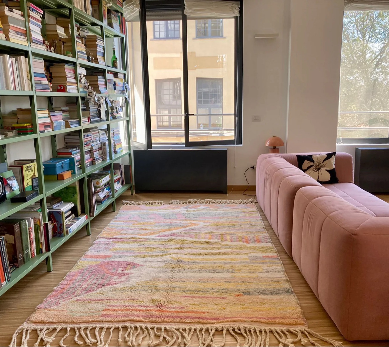 Large open plan living space with large windows, a wall of green shelving, pink pastel sofa and pastel coloured rug