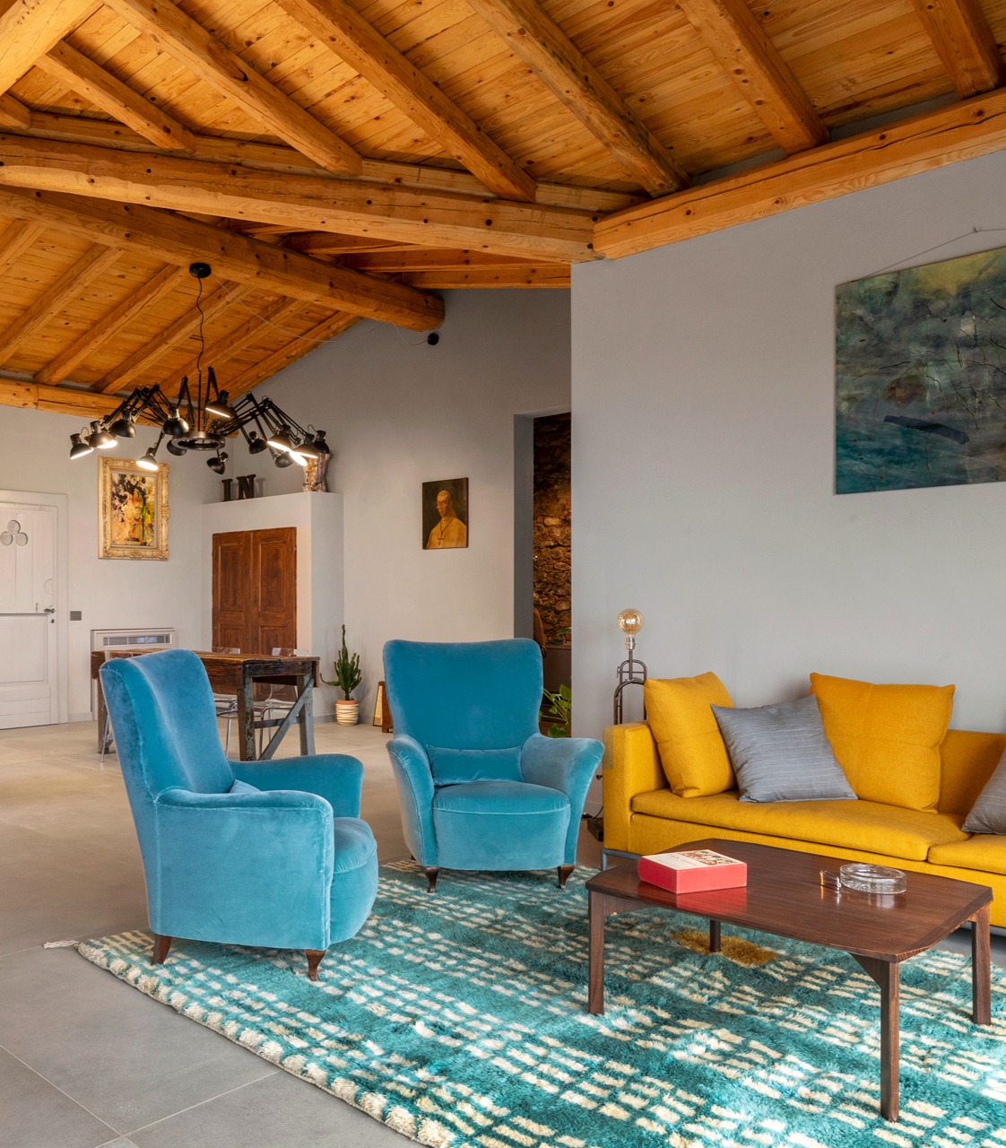 Modern living space with high wooden ceiling, 2 blue armchairs, yellow sofa, turquoise rug and dark wood coffee table. Small dining table in the background