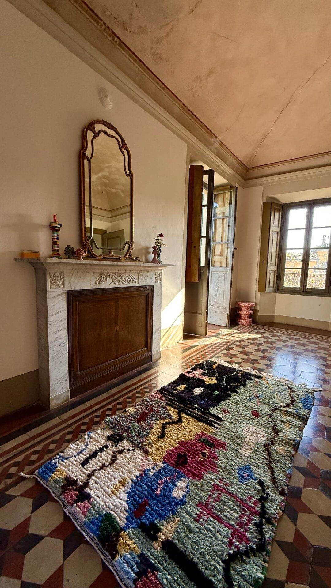 Grand living space with old fireplace, ornate doors and big window with shutters. Old tiled floor with multicoloured fluffy rug