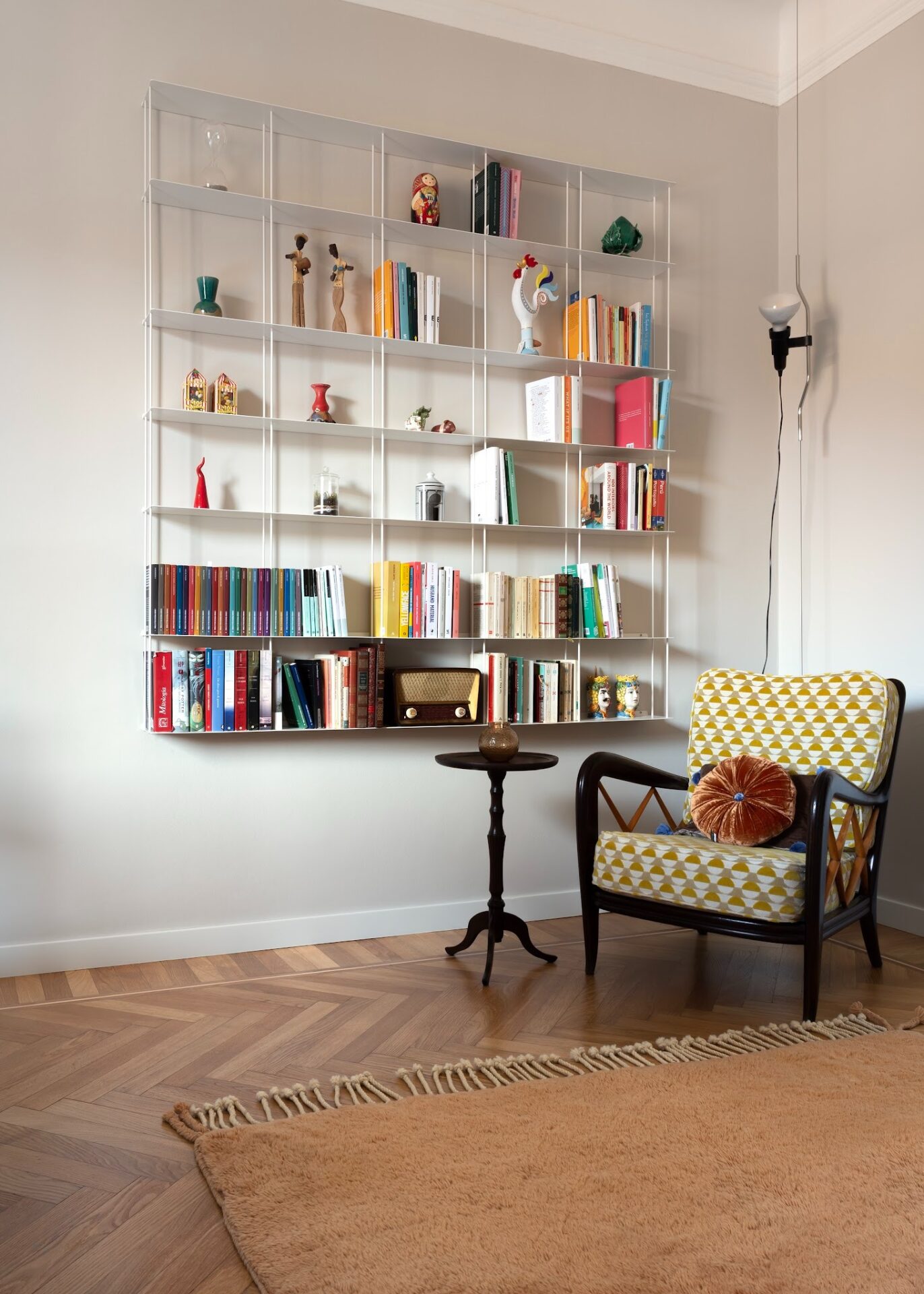 Corner of living room with wooden armchair and yellow cushions, small side table and white shelving with books. Wooden flooring with caramel rug