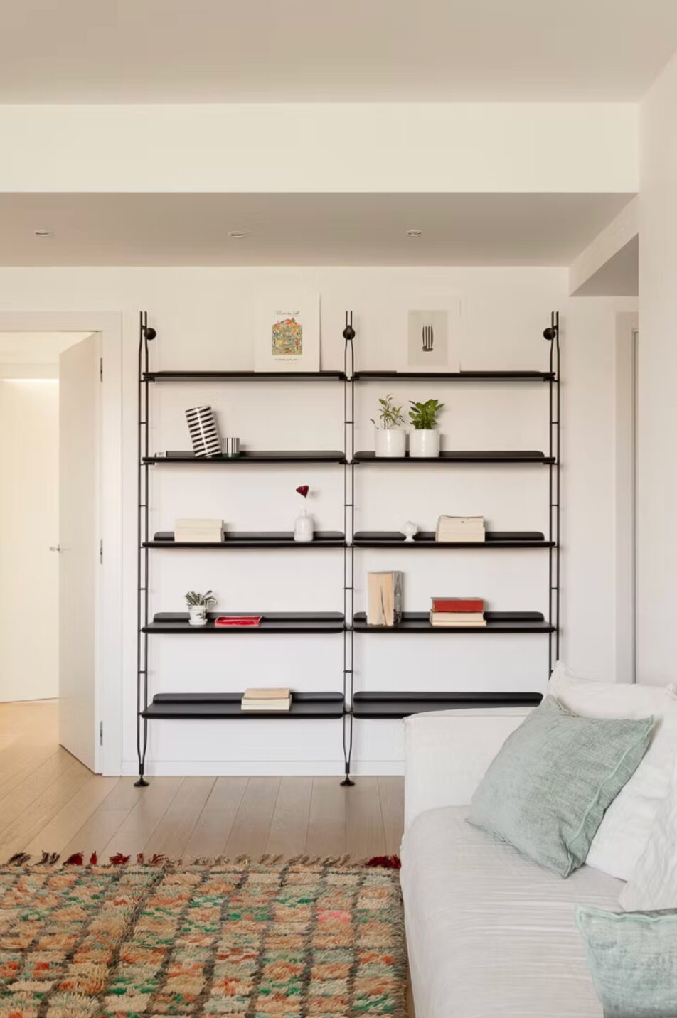 Modern simple living space with white walls, simple black and metal shelving - minimal ornaments and books. White sofa with pale blue cushions and a muted green statement rug.