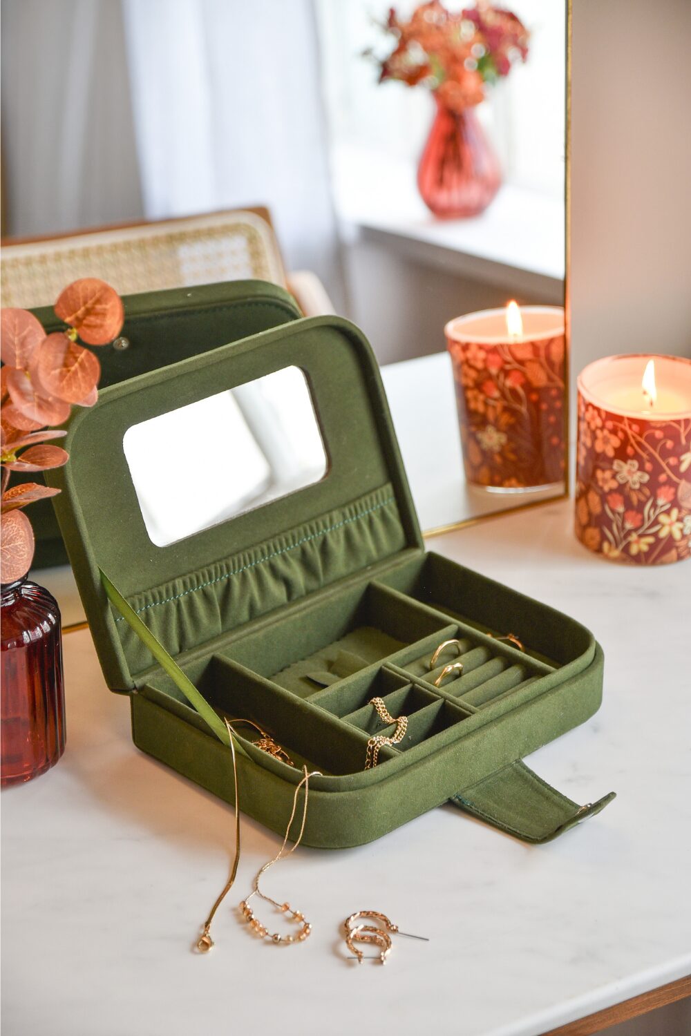 Green jewellery box with gold necklaces and rings on white dressing table next to orange candle and vase