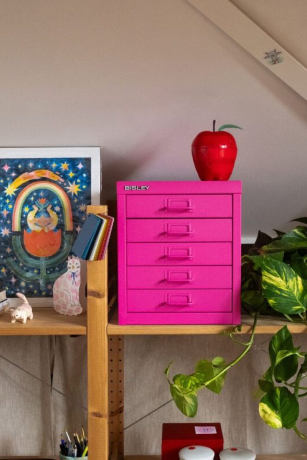 Bright pink metal drawers on wooden shelves holding pot plant, apple and painting