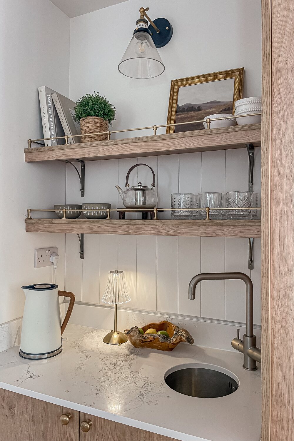 White kitchen counter with kettle, silver tap and gold lamp under wooden shelves of glassware and books