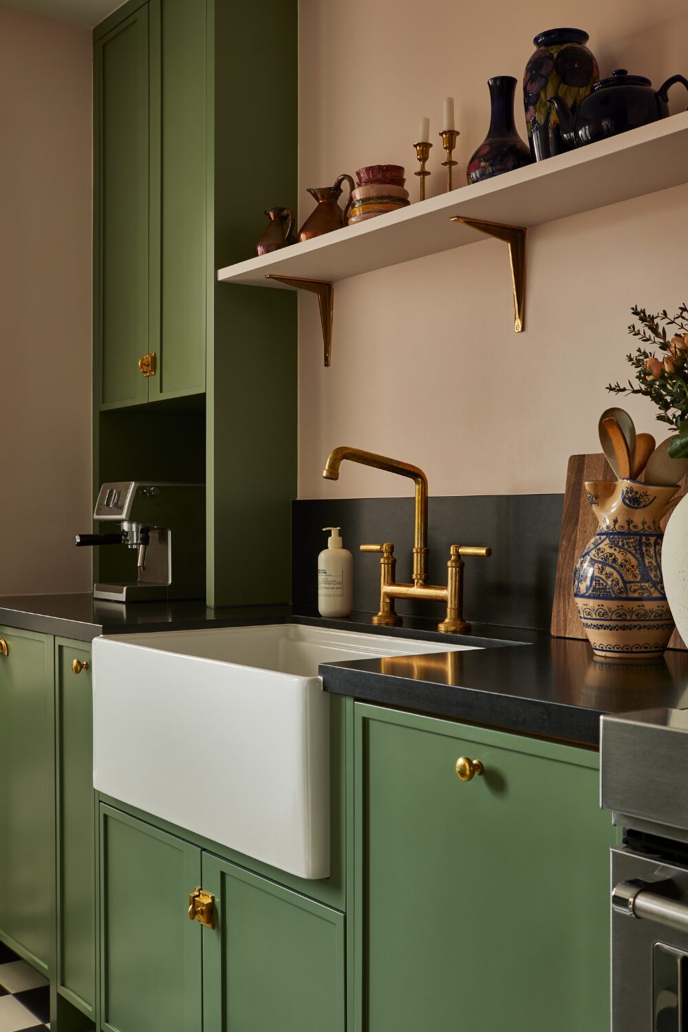Kitchen with dark green cabinets, and sink with gold taps under shelf of candlesticks and vases