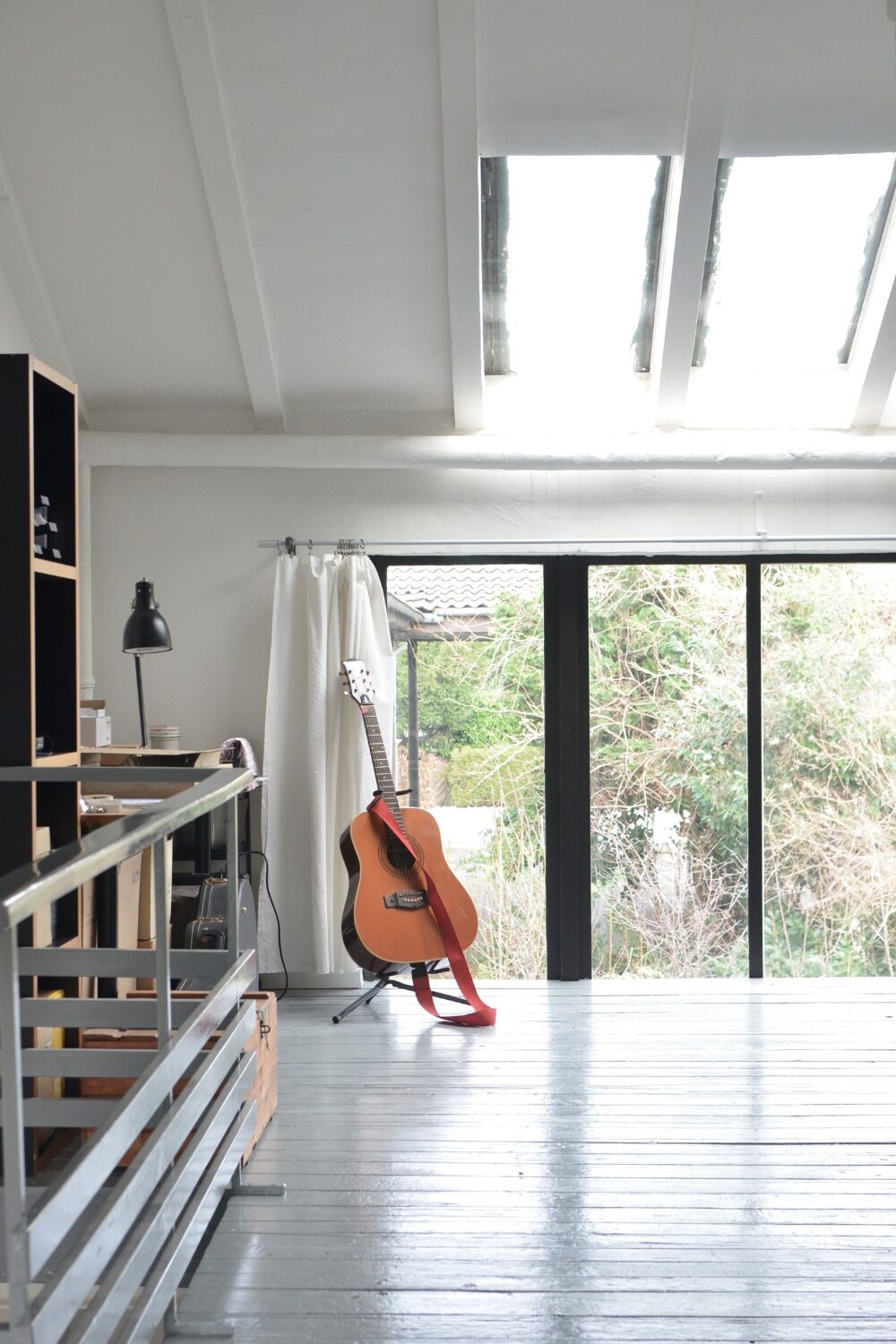 Room with grey wood floor, guitar on stand, desk, bookshelves and glass doors with garden view