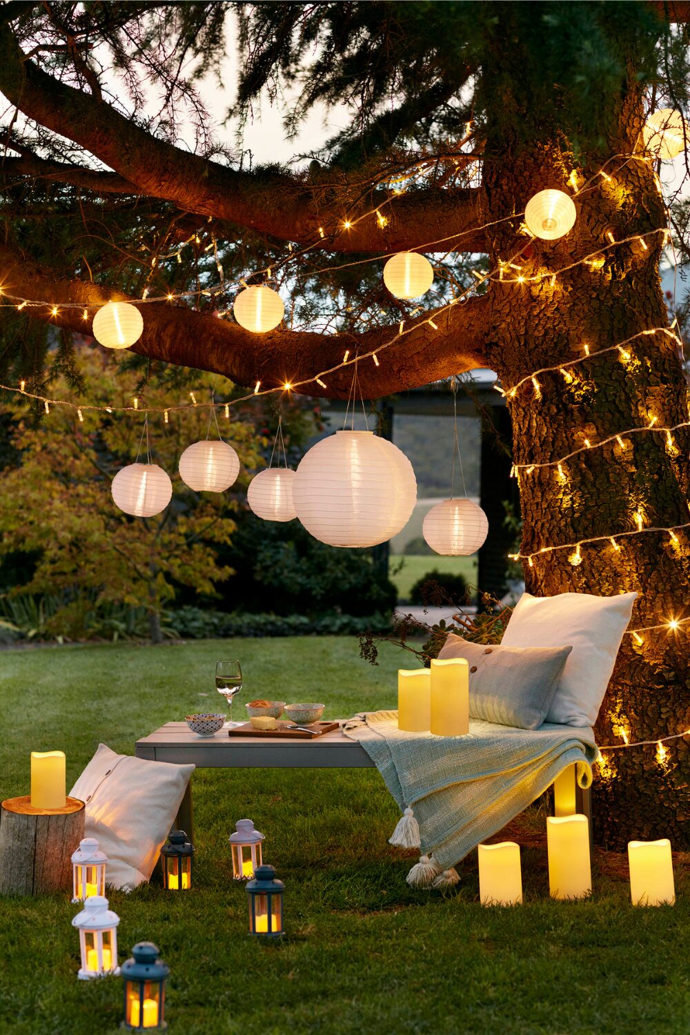 Wooden table on lawn under tree wrapped in lights, under paper lanterns and surrounded by candles