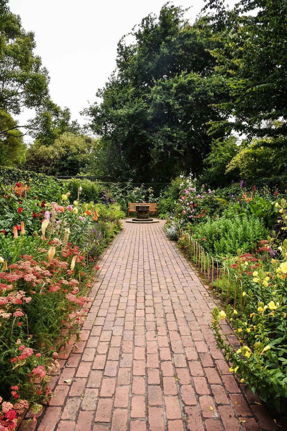 Lush flowered garden with brick path leading to stone fountain and wooden bench