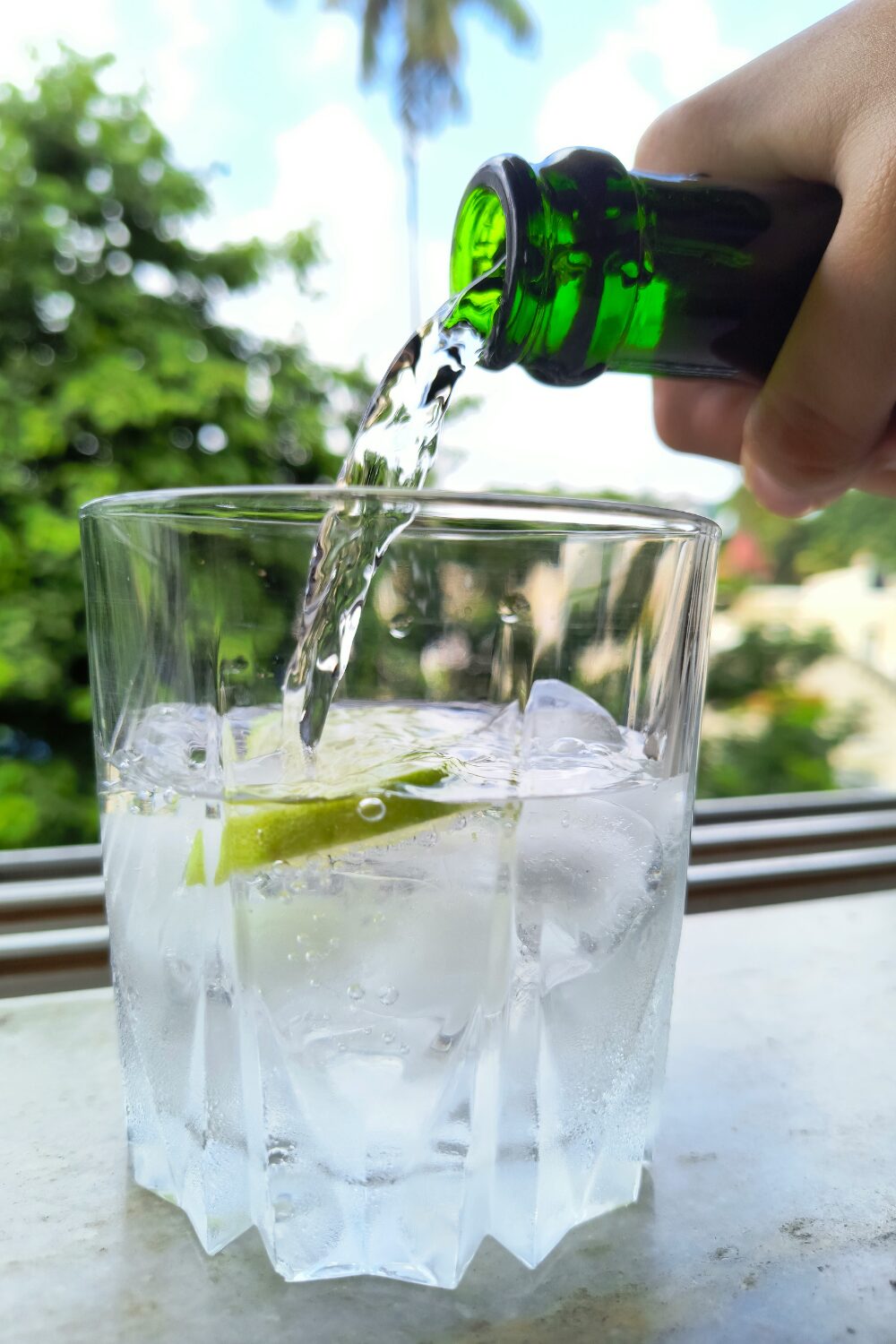 Hand pouring water from a green bottle into glass with ice and lime on windowsill