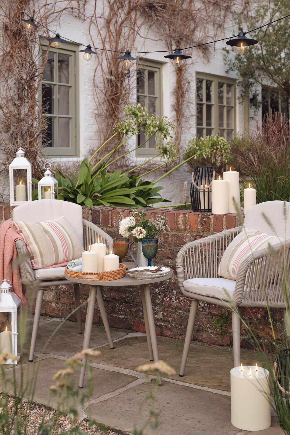 Two grey chairs and table on patio surrounded by white candles in front of white house with vines