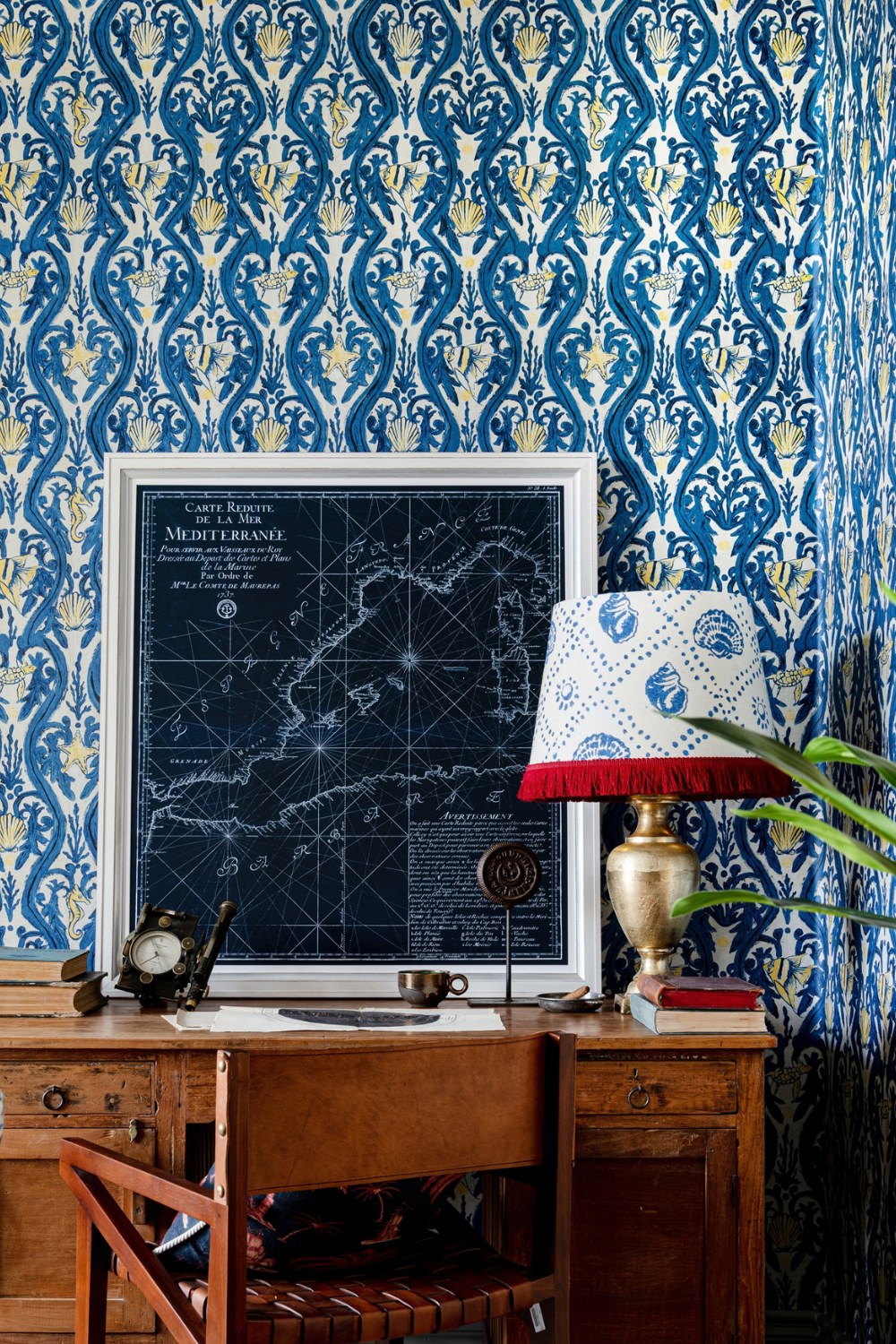 Framed map print resting on wooden desk next to lamp with fringed shade in room with blue print wallpaper