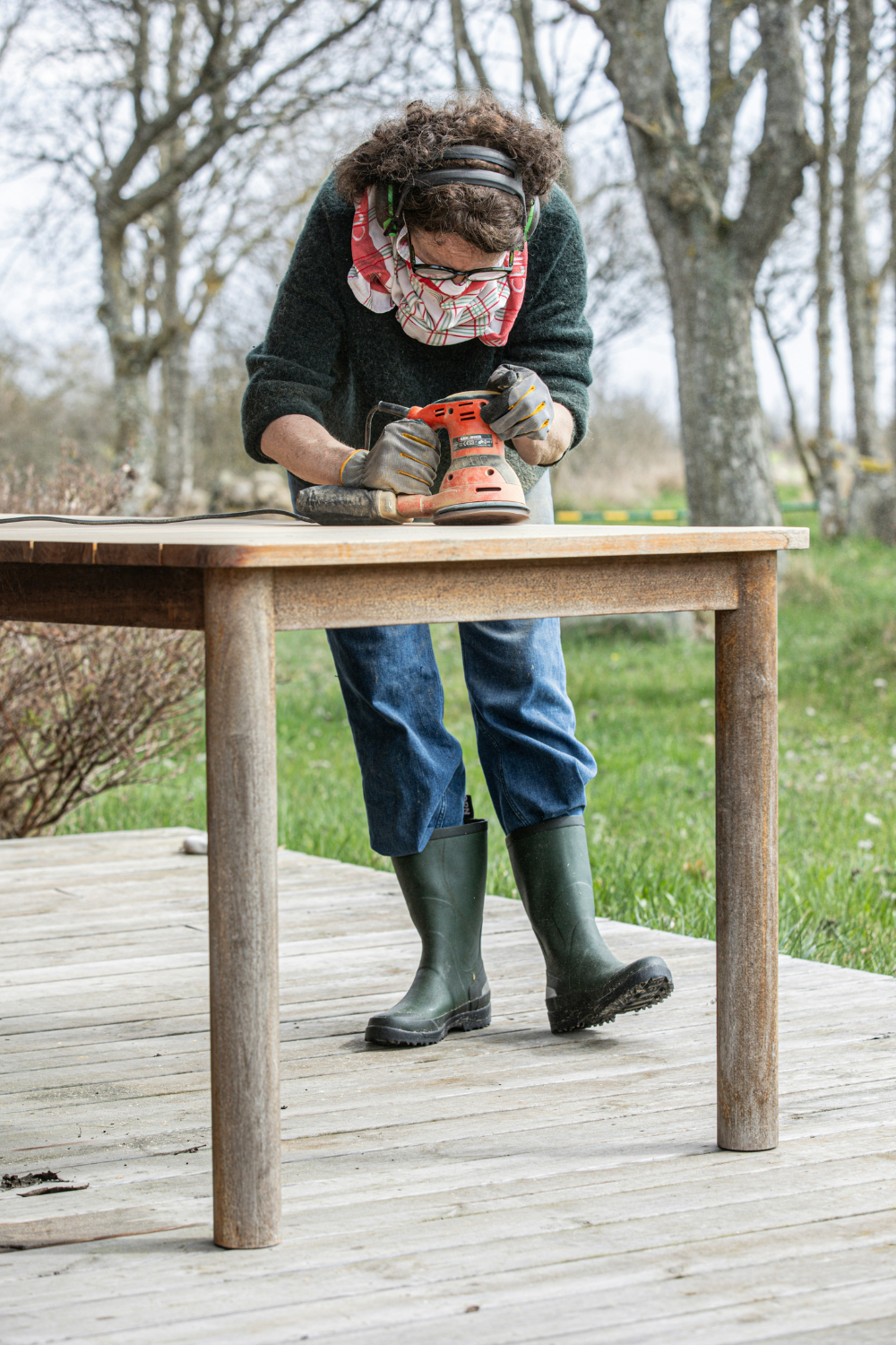 Dark-haired woman wearing jeans and wellies uses a tool to smooth a wooden table on a wooden deck with trees in background