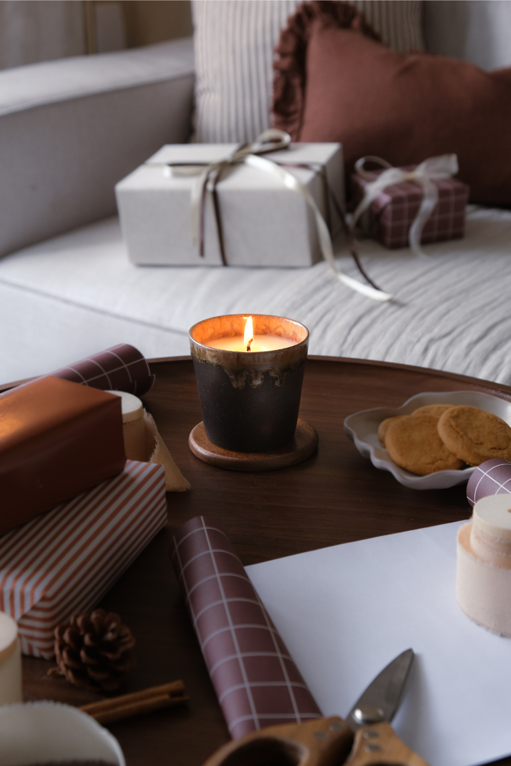 Jar candle on round wooden table next to plate of biscuits, wrapping paper, scissors, and presents