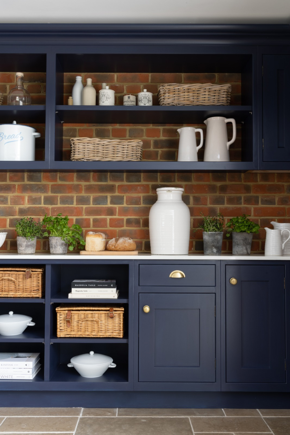 Navy kitchen cabinets and shelving holding white jugs and wicker baskets against brick wall