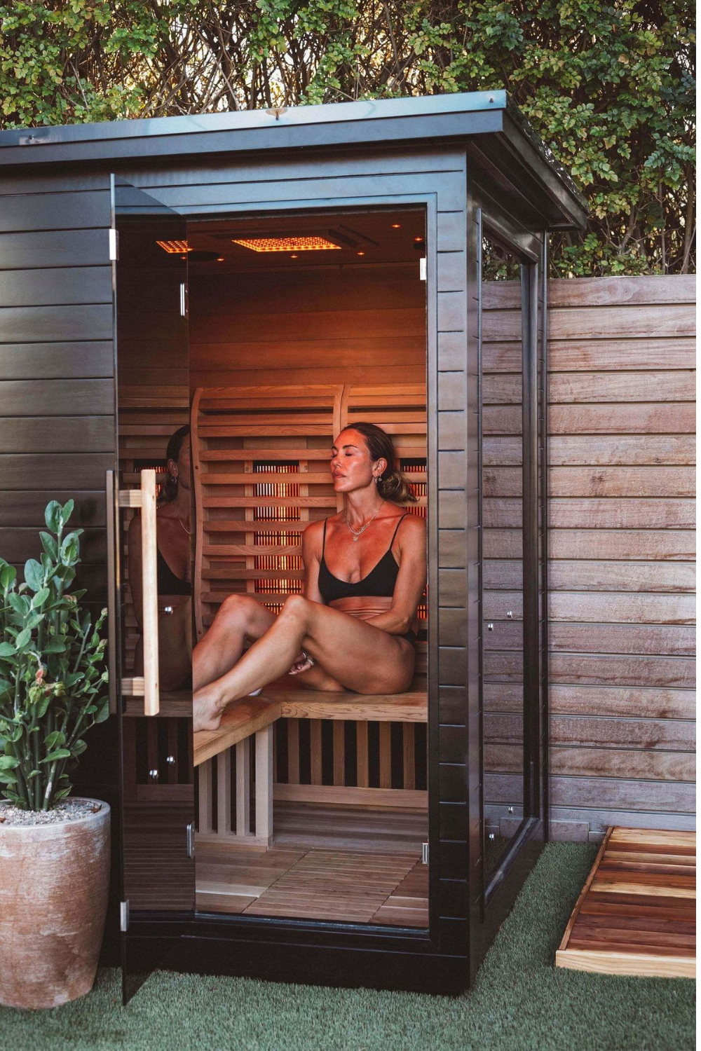 Woman sitting in small wooden sauna on lawn next to plant pot